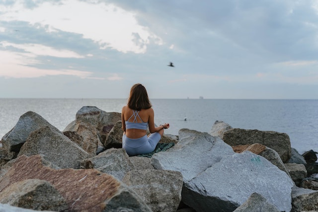 A woman in blue sportswear meditating on rocks near the sea.